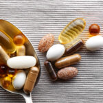 Close-up tablespoon filled with various dietary supplements, tablets and vitamins on a gray fabric background