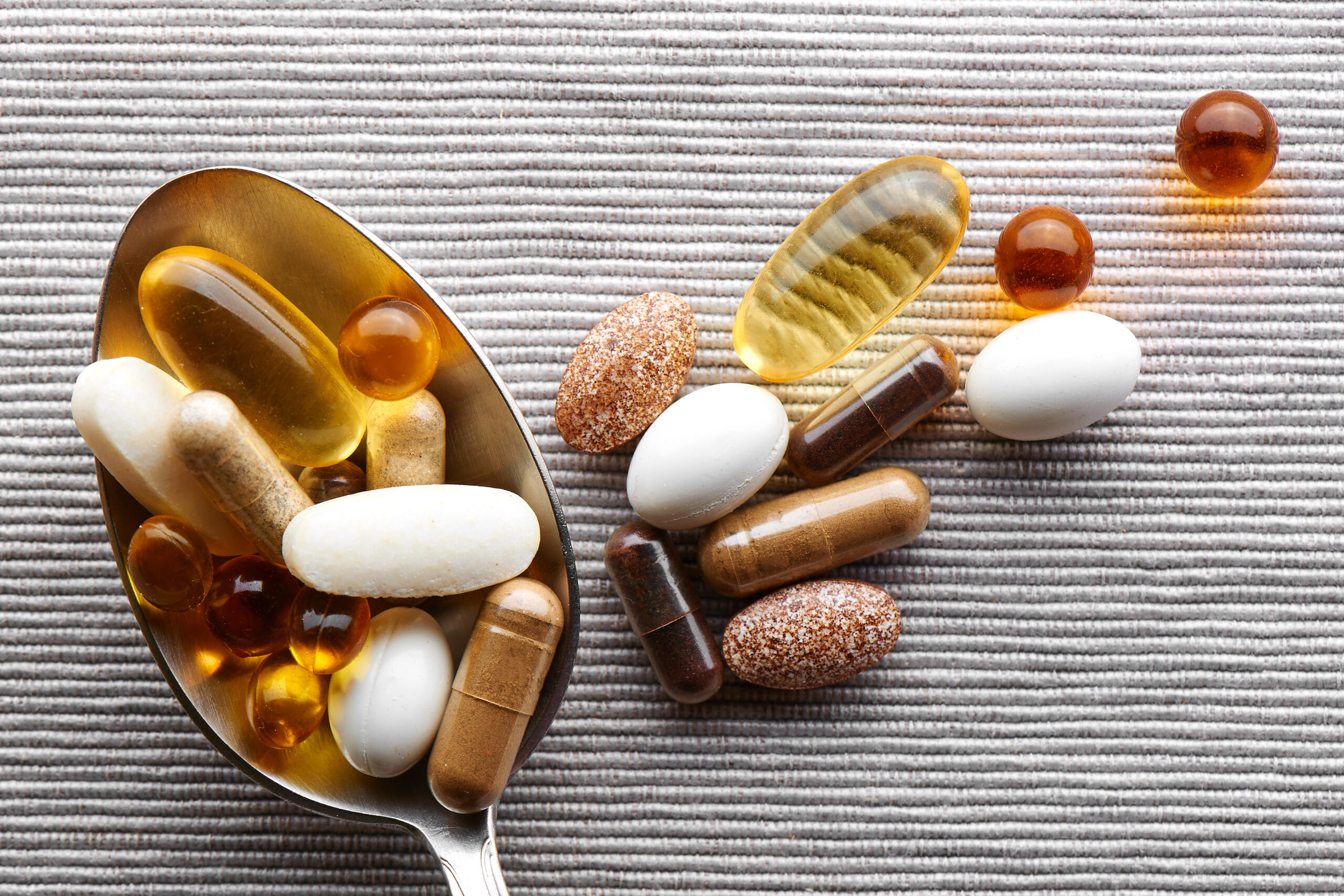 Close-up tablespoon filled with various dietary supplements, tablets and vitamins on a gray fabric background