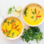 Two bowls of fresh coconut curry soup with cut green parsley, slices of lime, spoons, isolated over white background. Curry bowl for dinner. Vegetarian food concept