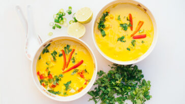 Two bowls of fresh coconut curry soup with cut green parsley, slices of lime, spoons, isolated over white background. Curry bowl for dinner. Vegetarian food concept