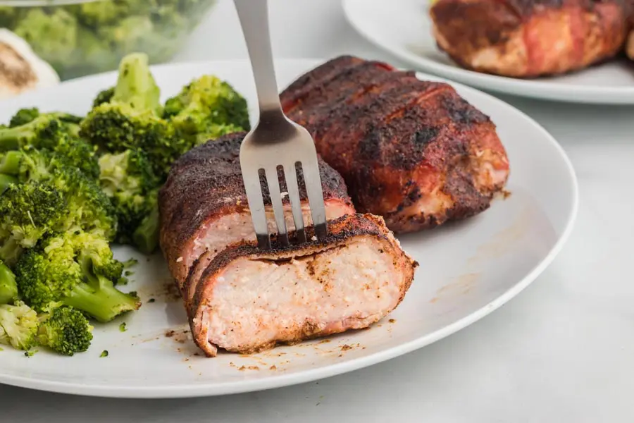 A fork going through a slice of cooked pork chops wrapped in bacon. Juices come out of the pork chop.